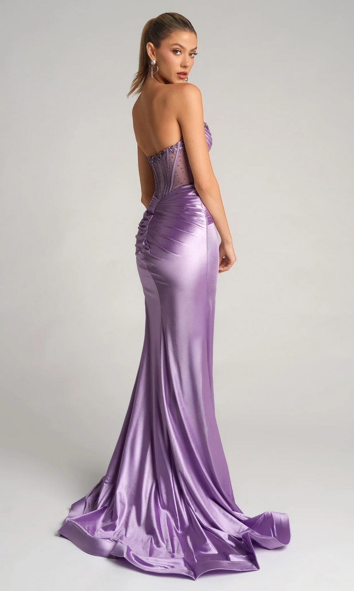 A woman in the Portia and Scarlett PS26085, a strapless lavender formal gown with an embellished corset bodice, sheer back panel, satin skirt, and ruched details, stands against a plain background.